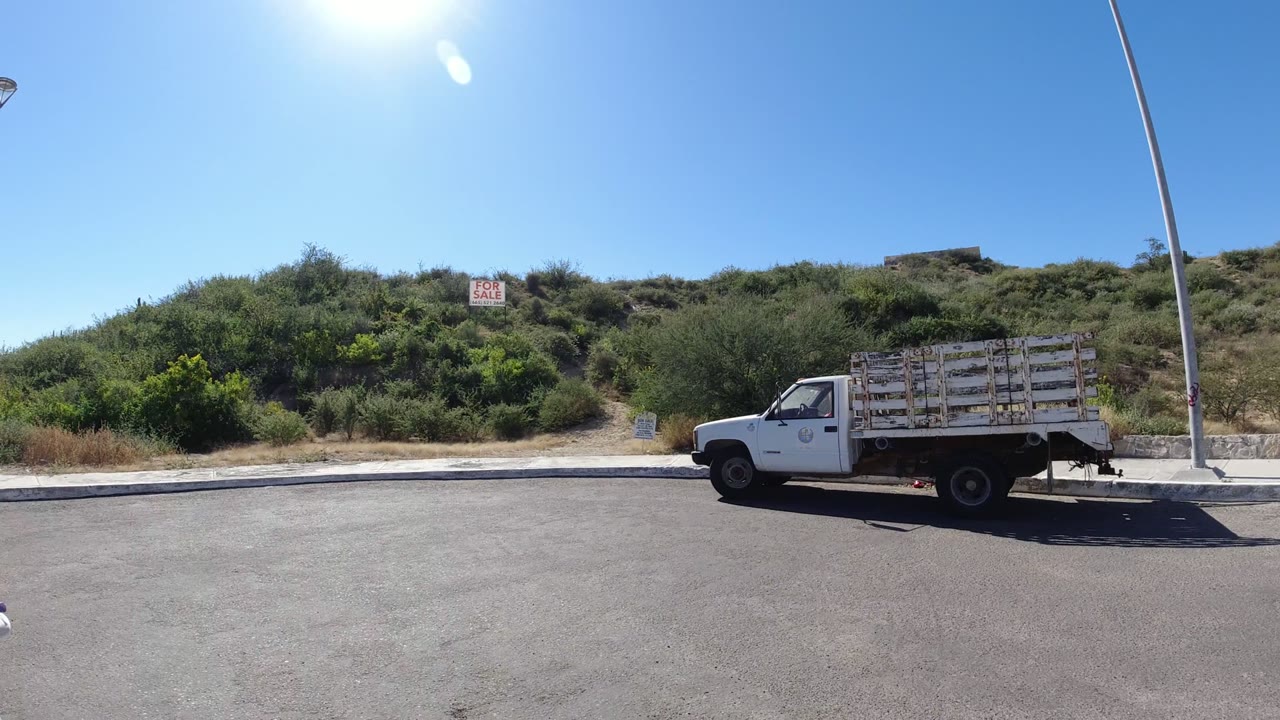 Very wide San Jose del Cabo street view with a parked white truck, dry hillside vegetation, small roadside signs, a tall streetlight, and no visible people.