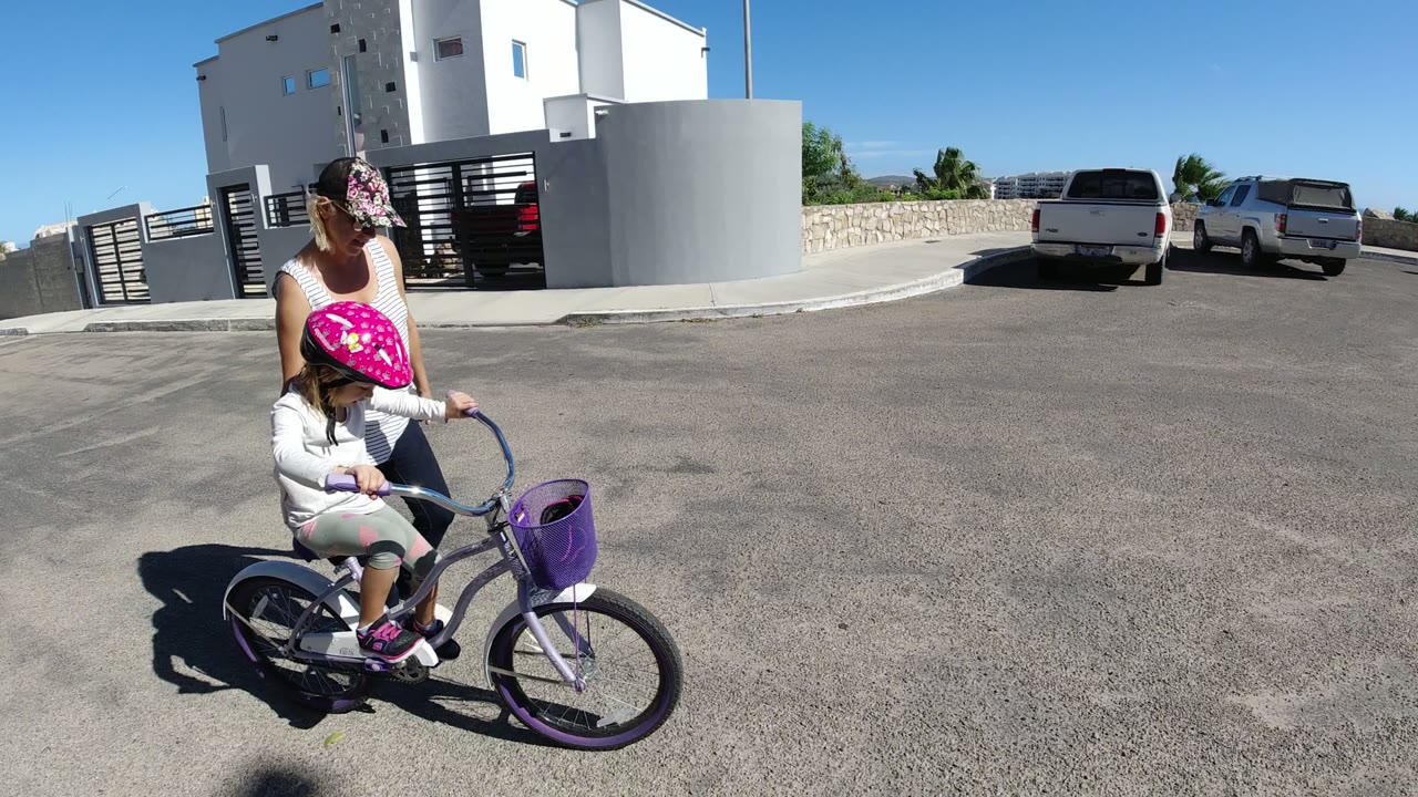 Adult standing beside a child on a small purple bicycle on a sunny paved street.