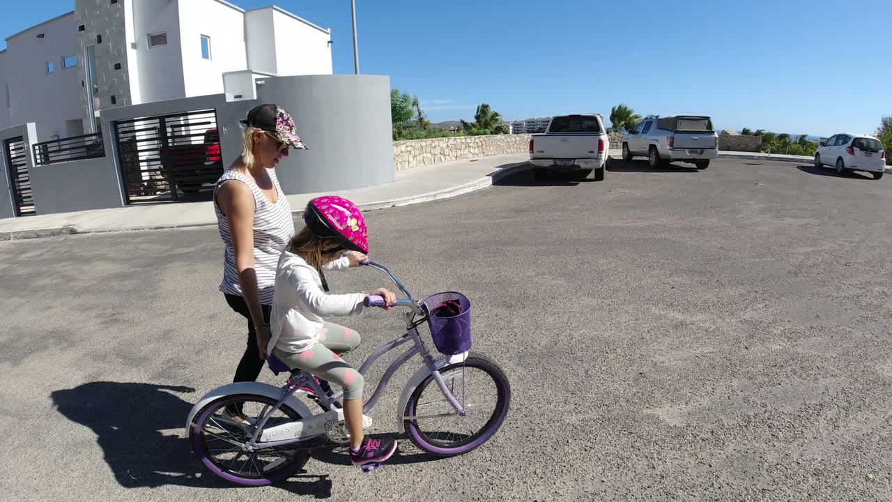The child pedals the purple bicycle as the adult walks at her side, with white buildings, a gate, pickup trucks, and bright sun visible in San Jose del Cabo, Mexico.