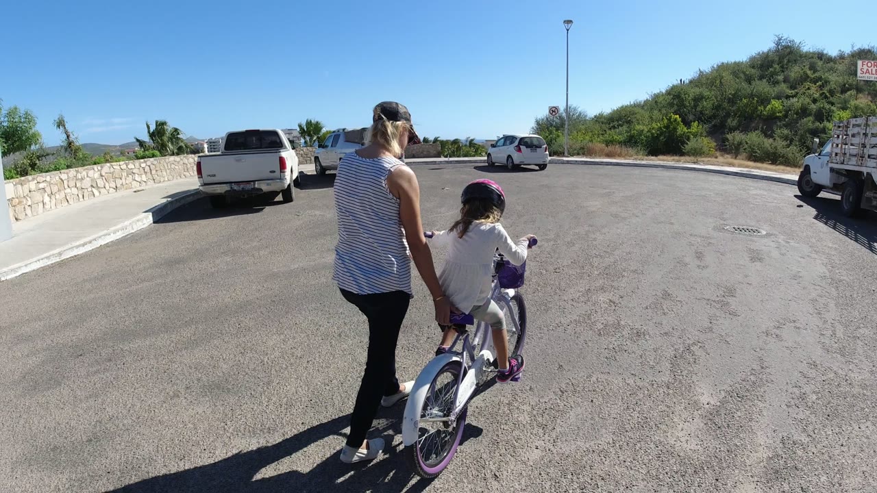 Adult helping a child on a small bicycle in a sunny parking area.