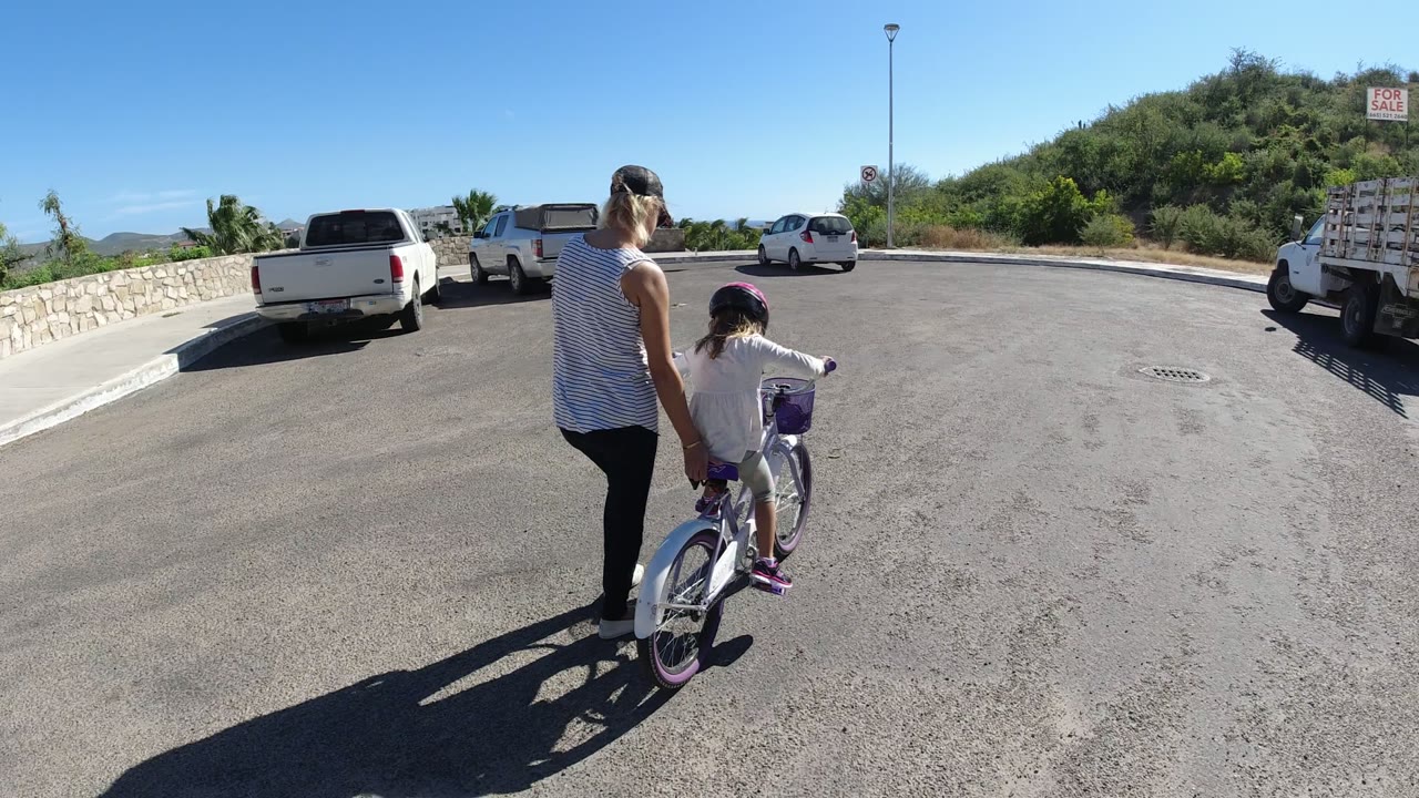 The child pedals farther into the parking area while the adult stays close on the left, with two pickup trucks, a white car, curb, hillside, and clear sky in view in San Jose del Cabo.