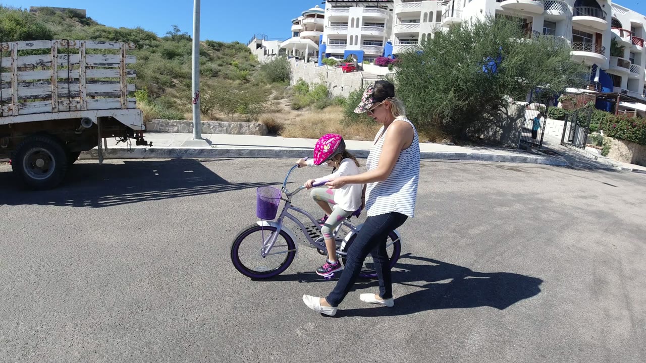 Woman walking beside a girl on a purple bicycle on a sunny road.