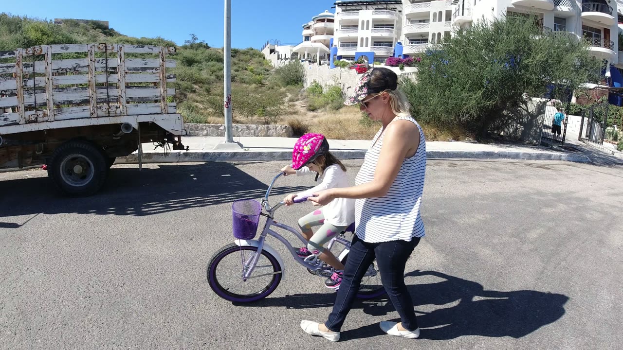 The girl rides farther across the road in San Jose del Cabo, Mexico, with the woman close at her side and a parked utility truck behind them.