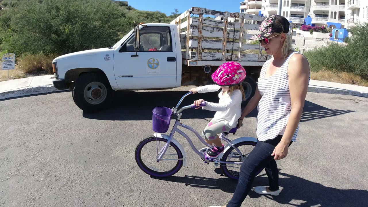 Mother walking beside a young girl on a purple bicycle near a white truck.