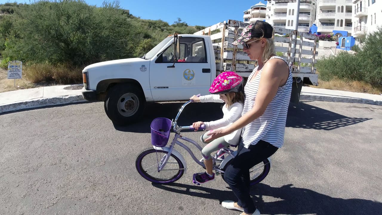 A helmeted child pedals a purple bike while her mother walks close at her side, with a parked white truck and hillside buildings behind them in San Jose Del Cabo, Mexico.