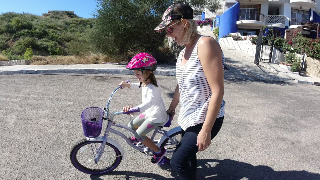 In San Jose del Cabo, a woman walks next to a young girl in a pink helmet as the child rides a small bicycle with a purple basket along a paved parking area.