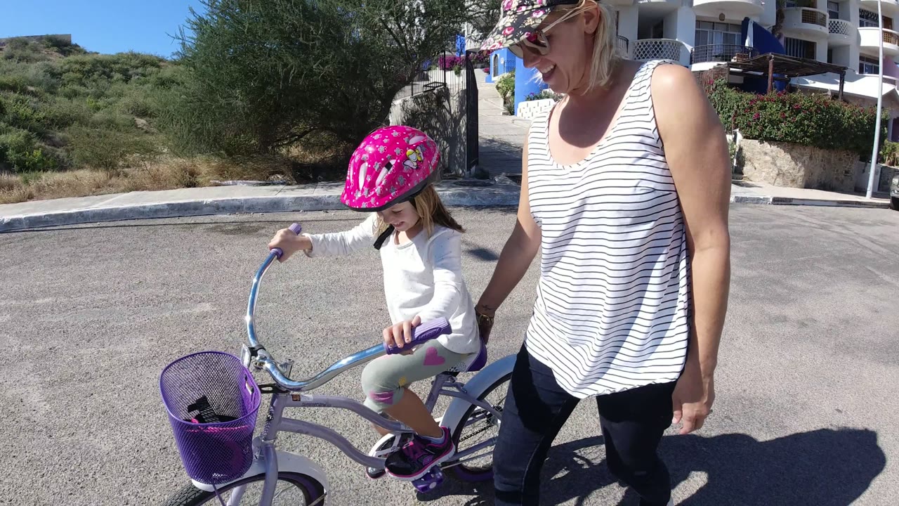 The girl rides beside the woman with both hands on the handlebars, with white buildings, balconies, shrubs, and a curb visible behind them in San Jose del Cabo.