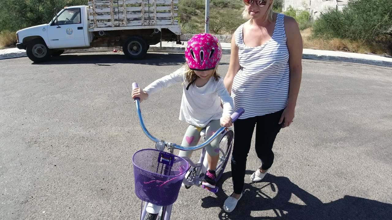At 0:00 in San Jose del Cabo, a child in a bright pink helmet grips a purple bicycle while an adult woman stands beside her on a sunlit residential street.