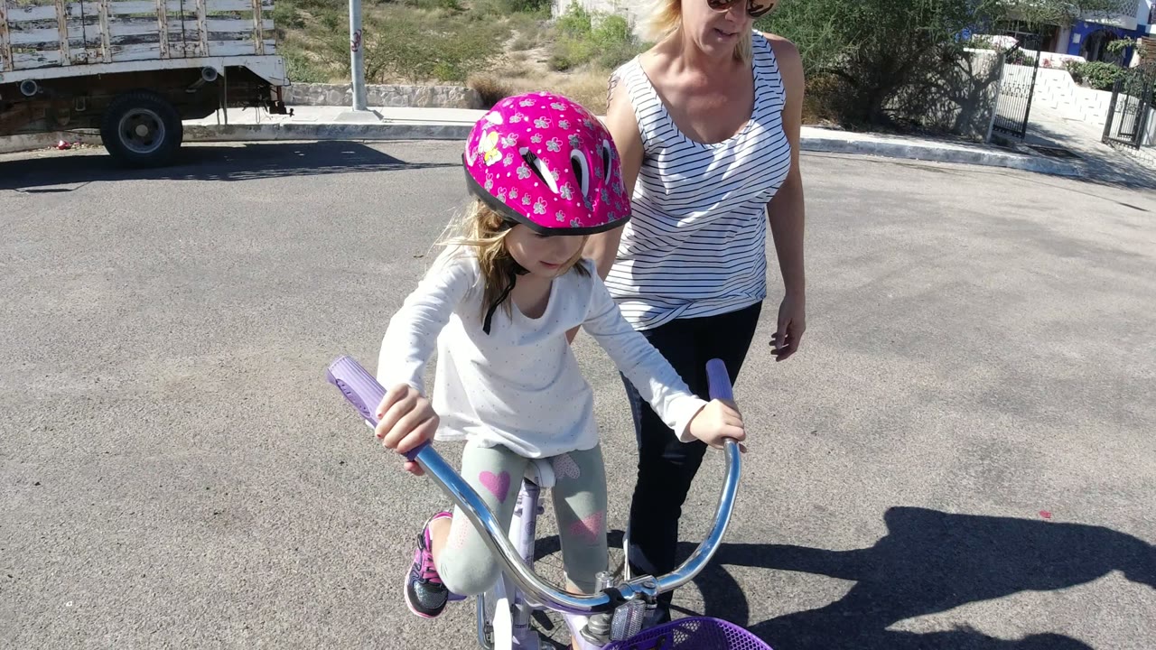 At 0:01 in San Jose del Cabo, the girl leans over the handlebars of her purple and white bicycle as the woman walks near her shoulder in bright sun.