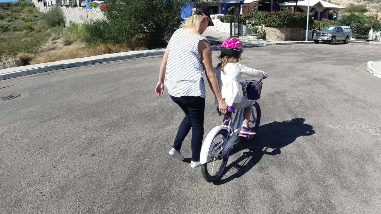 In San Jose del Cabo, Mexico, the child pedals the purple-and-white bicycle while the woman walks just to the left, with open road ahead and homes beyond.