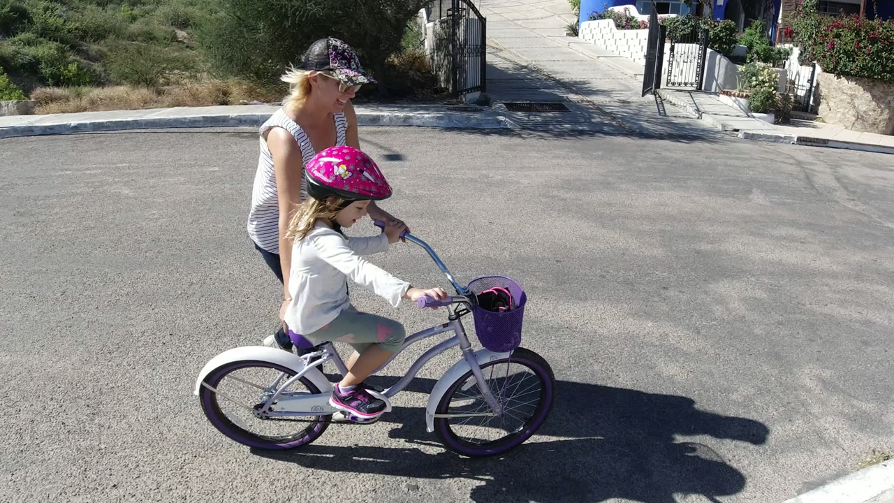 Woman walking beside a helmeted girl on a small bicycle in a sunlit street.