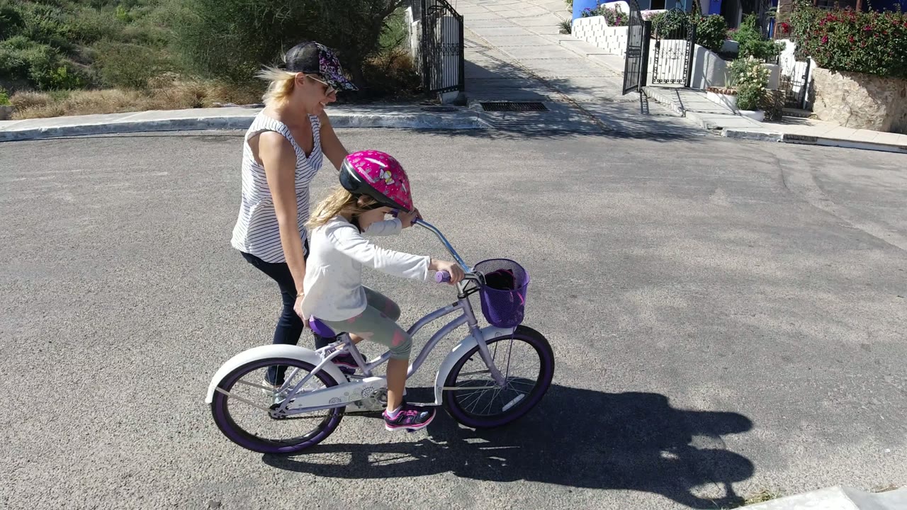The girl continues riding the small white bicycle in San Jose del Cabo while the woman stays just behind her shoulder on the open street.
