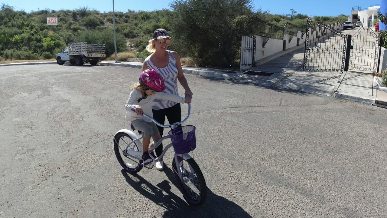 Woman guiding a child on a bicycle in a sunny paved street.