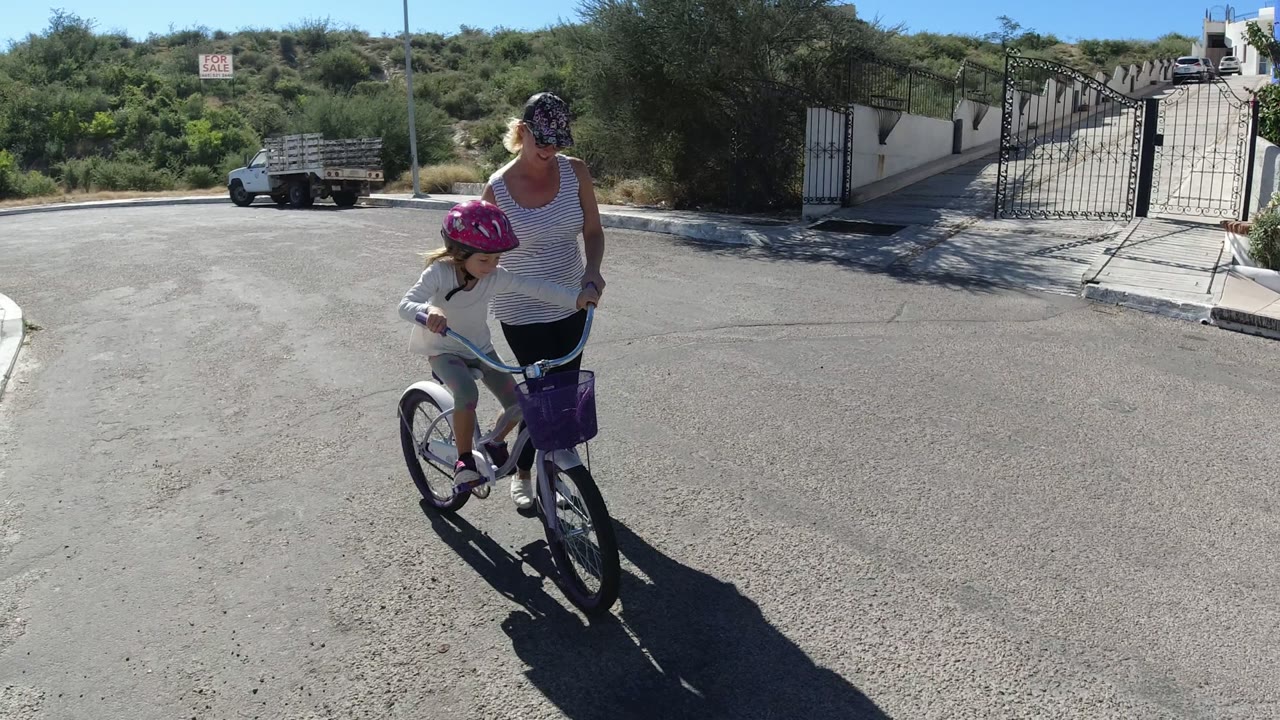 One second in, the child pedals slightly ahead of the woman on the sunlit San Jose del Cabo street, with the purple basket centered and the open gate behind them.