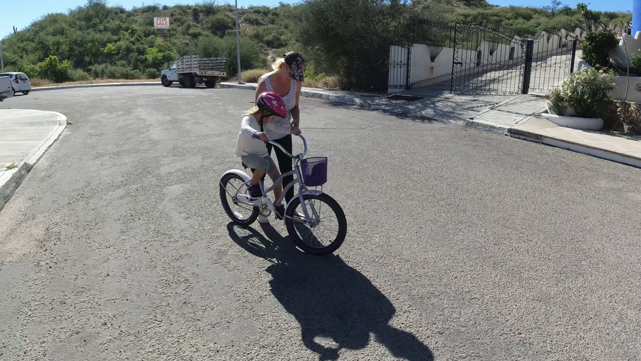 One second in, the child remains on the bicycle while the woman stays alongside, with a gated driveway, hillside vegetation, and a small white car visible behind them in San Jose Del Cabo.