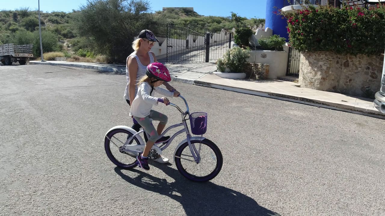 Woman walking beside a child on a small bicycle in a sunny street.
