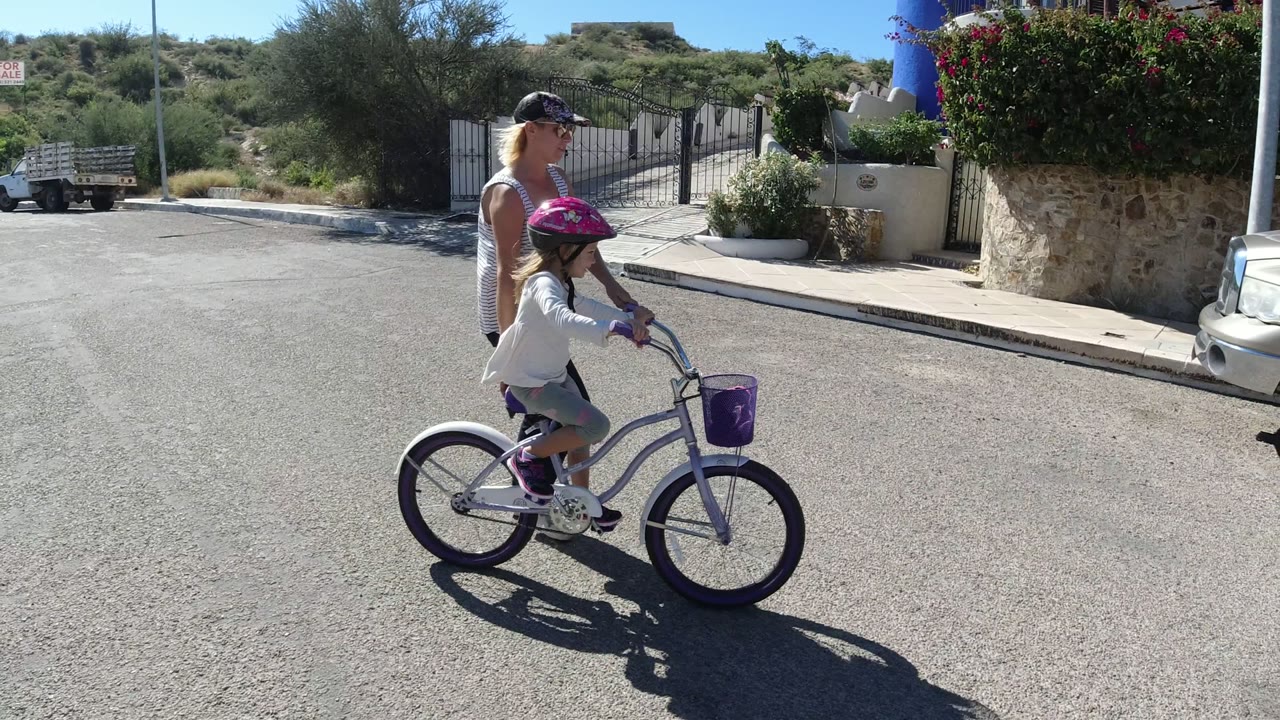 The child pedals the small bicycle forward while the woman stays close behind, with a stone wall, metal gate, and sunlit roadside plants behind them in San Jose del Cabo.