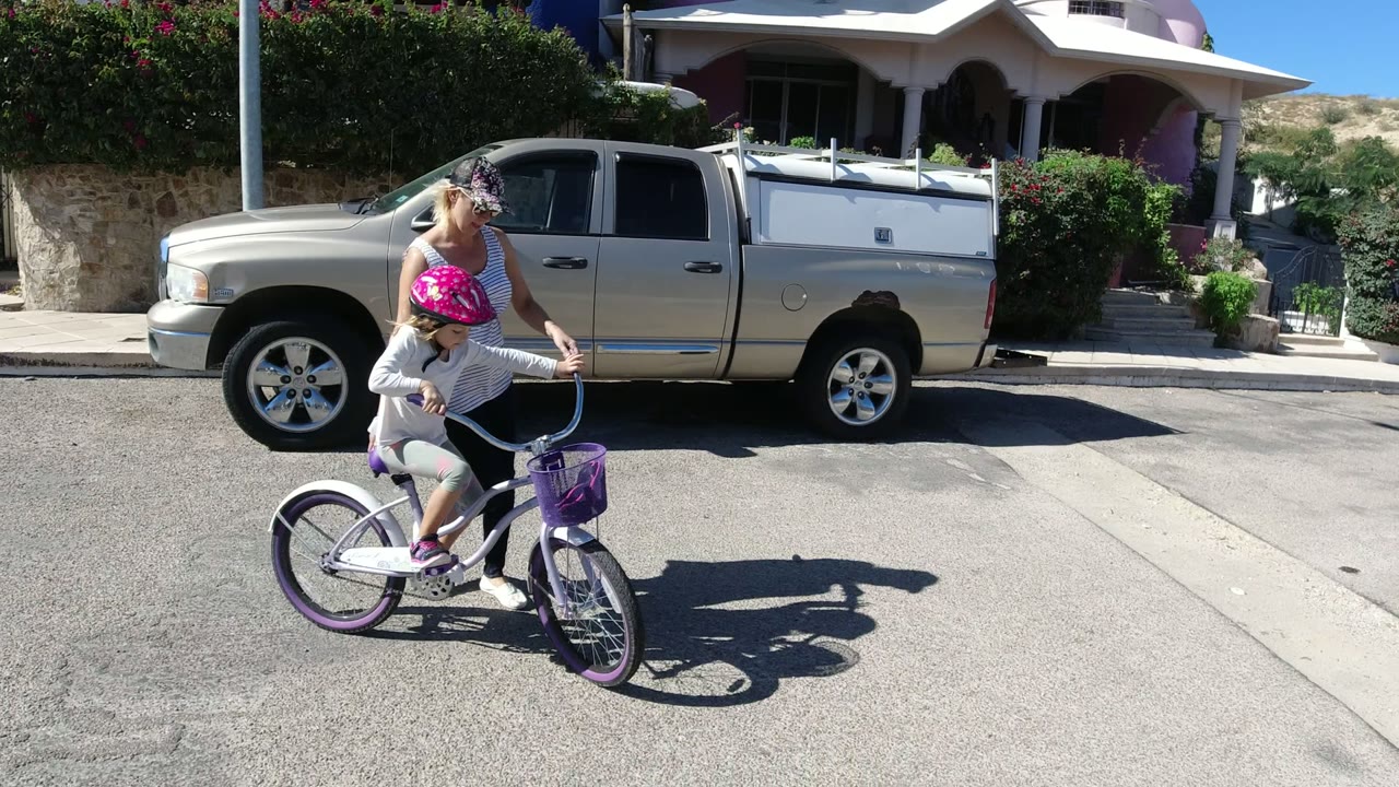 In San Jose del Cabo, Mexico, a young girl in a pink helmet rides a purple-and-white bicycle while her mother walks close beside her near a parked pickup and private home.