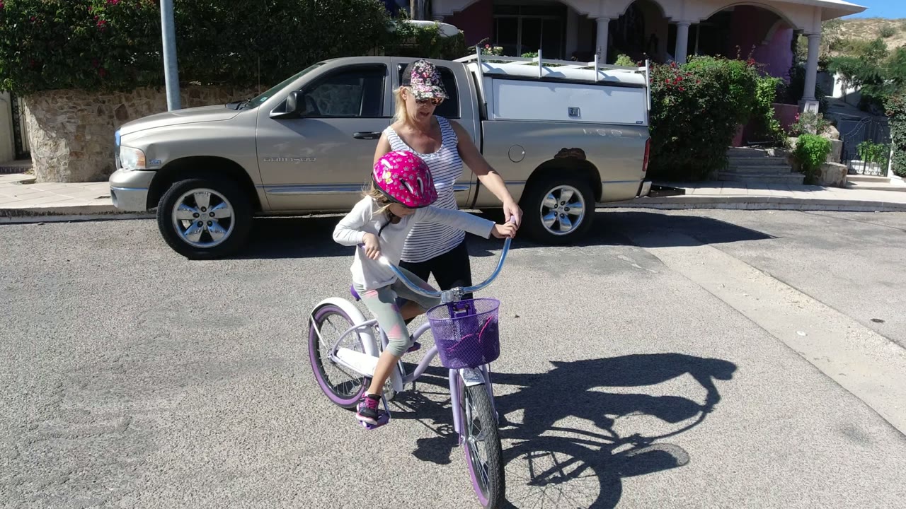 The girl pedals the small bicycle near the center of the street as her mother keeps one hand near the handlebars, with a pickup truck and residential entry behind them in San Jose del Cabo, Mexico.