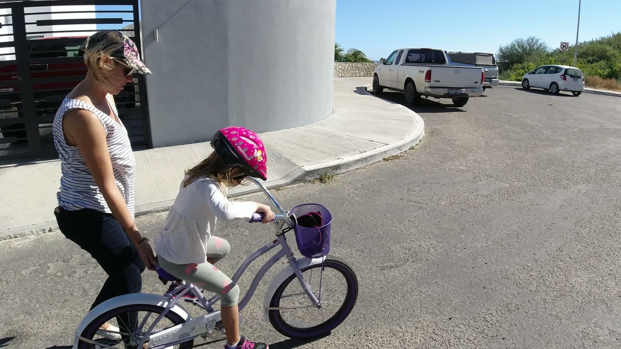 In San Jose del Cabo, a woman walks close behind a young girl on a purple bicycle with training wheels beside a white building column and paved driveway.
