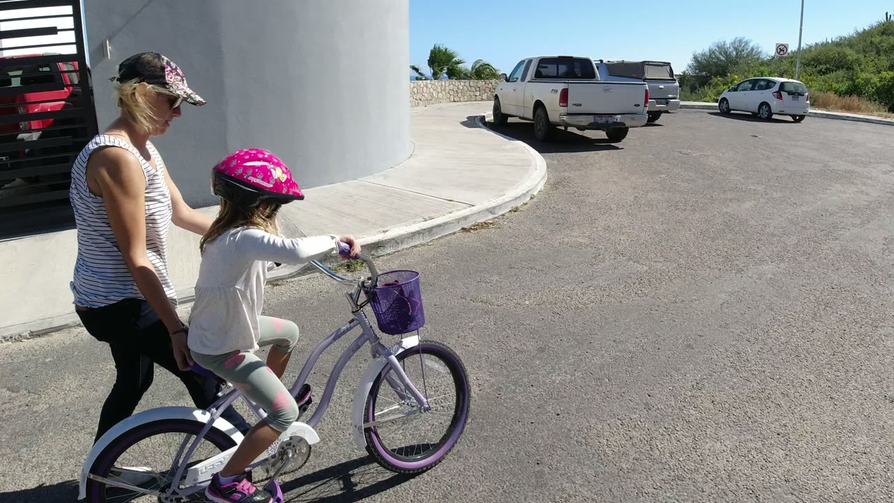 The child pedals the purple bike forward in San Jose del Cabo while the woman stays beside the rear wheel, with parked vehicles and a curved curb ahead.