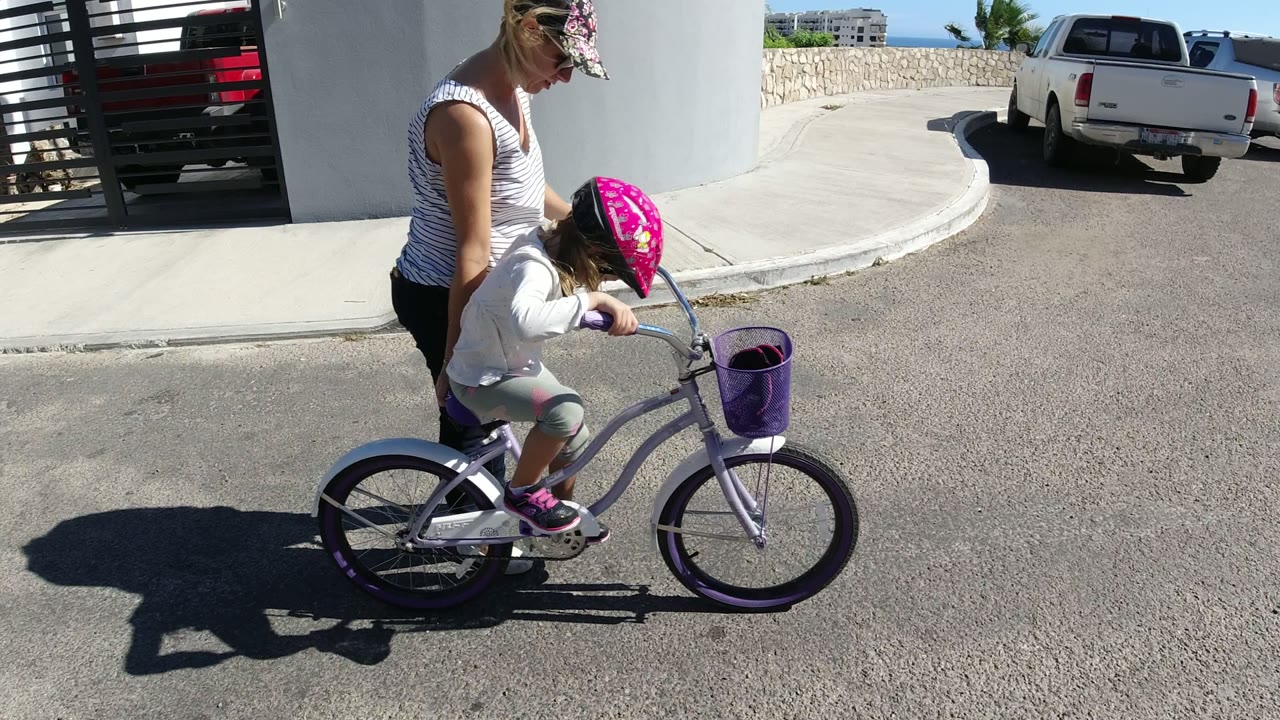 In San Jose del Cabo, a woman walks beside a young girl wearing a pink helmet as the child rides a purple bicycle near a gate, white wall, and parked pickup trucks.