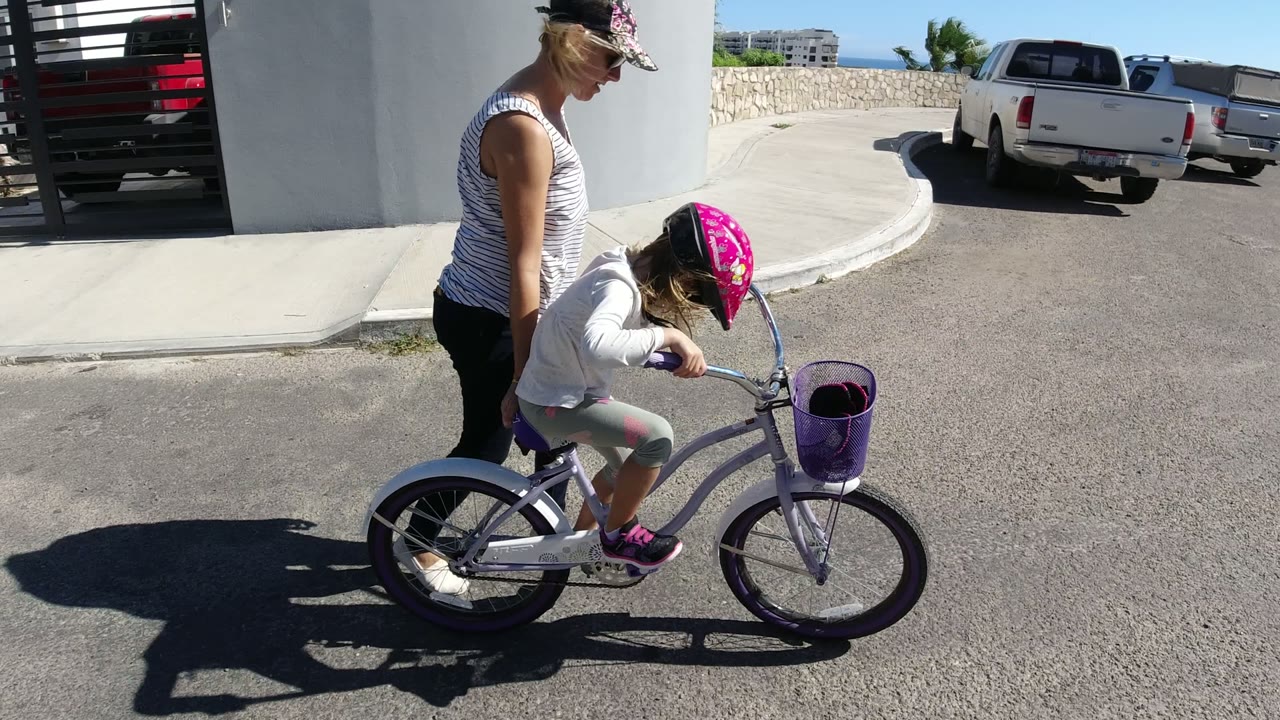 In San Jose del Cabo, the young girl pedals a purple bike while her mother stays just behind her, with a white pickup truck and low stone wall in the background.