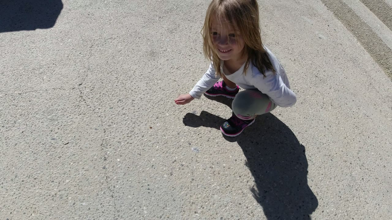At 0:01, the smiling girl is seen from a high angle on pale concrete in San Jose del Cabo, Mexico, with strong sun defining her hair, clothing, and shadow.