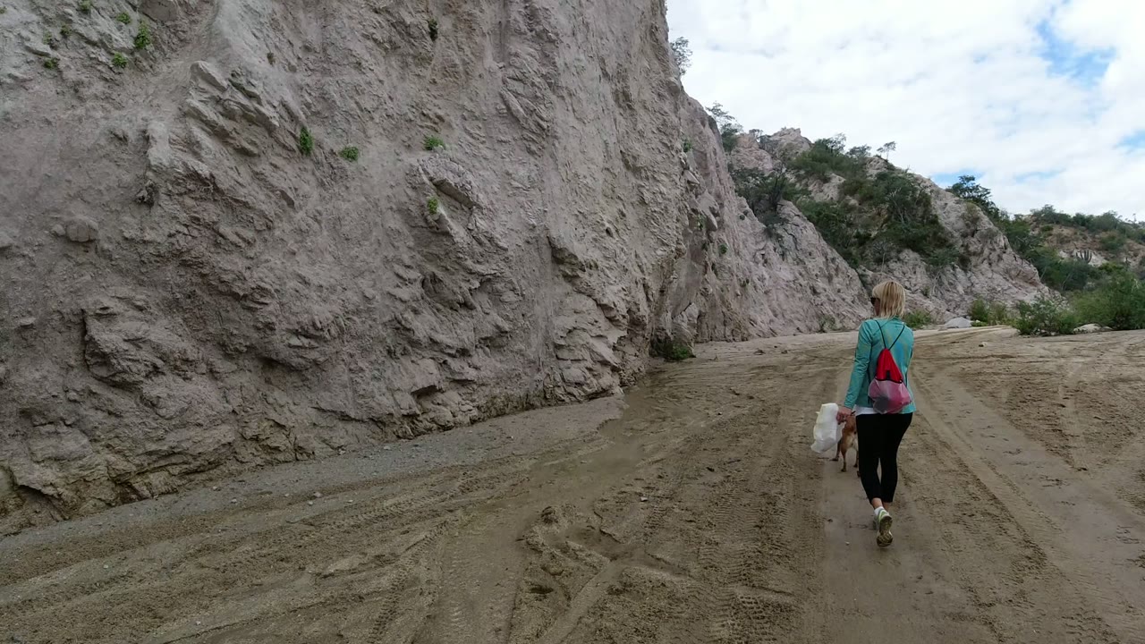 A woman walks away with a dog on a sandy trail between tall gray rock walls in San Jose del Cabo, Mexico, at the start of the clip.