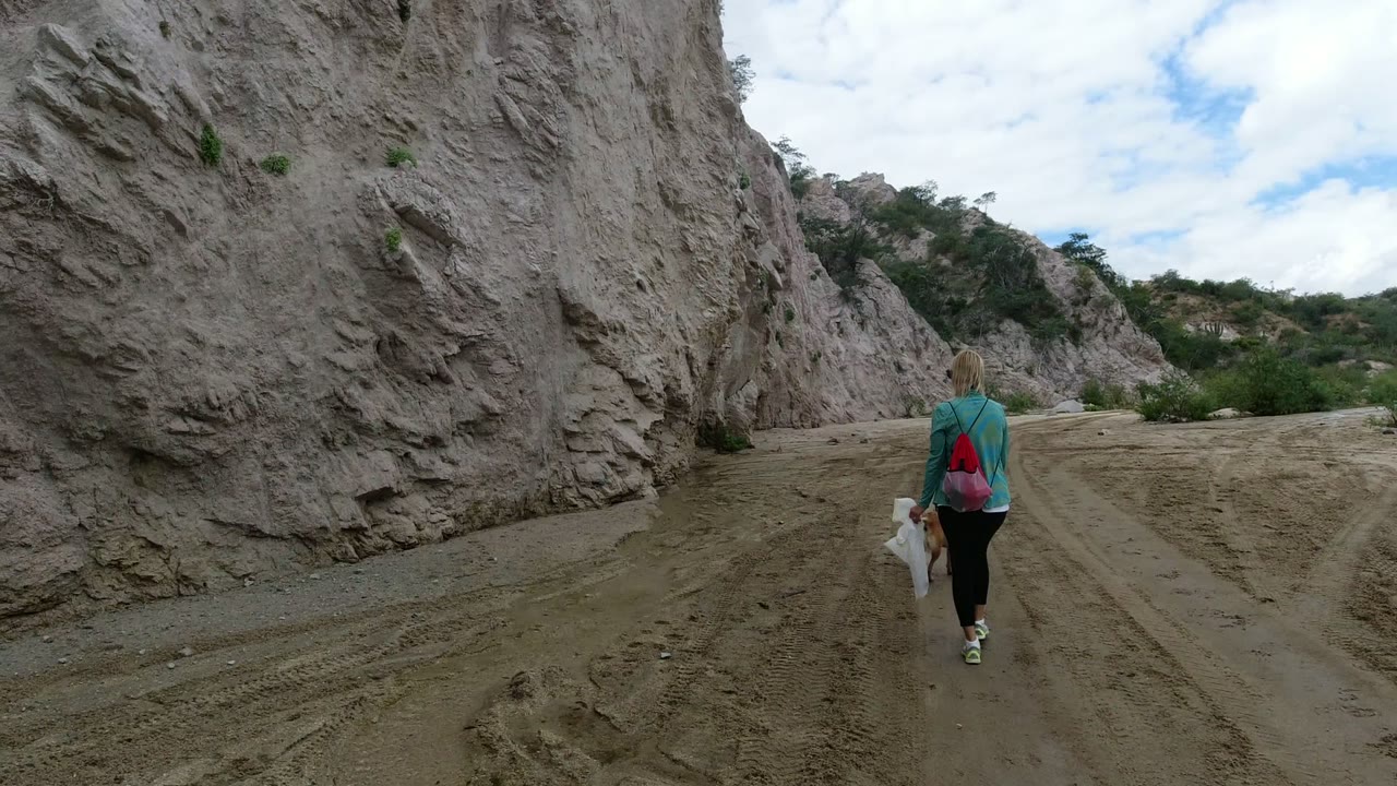 The woman and dog continue away from camera on the sandy trail in San Jose del Cabo, with the left rock wall close to frame and cloudy sky above.