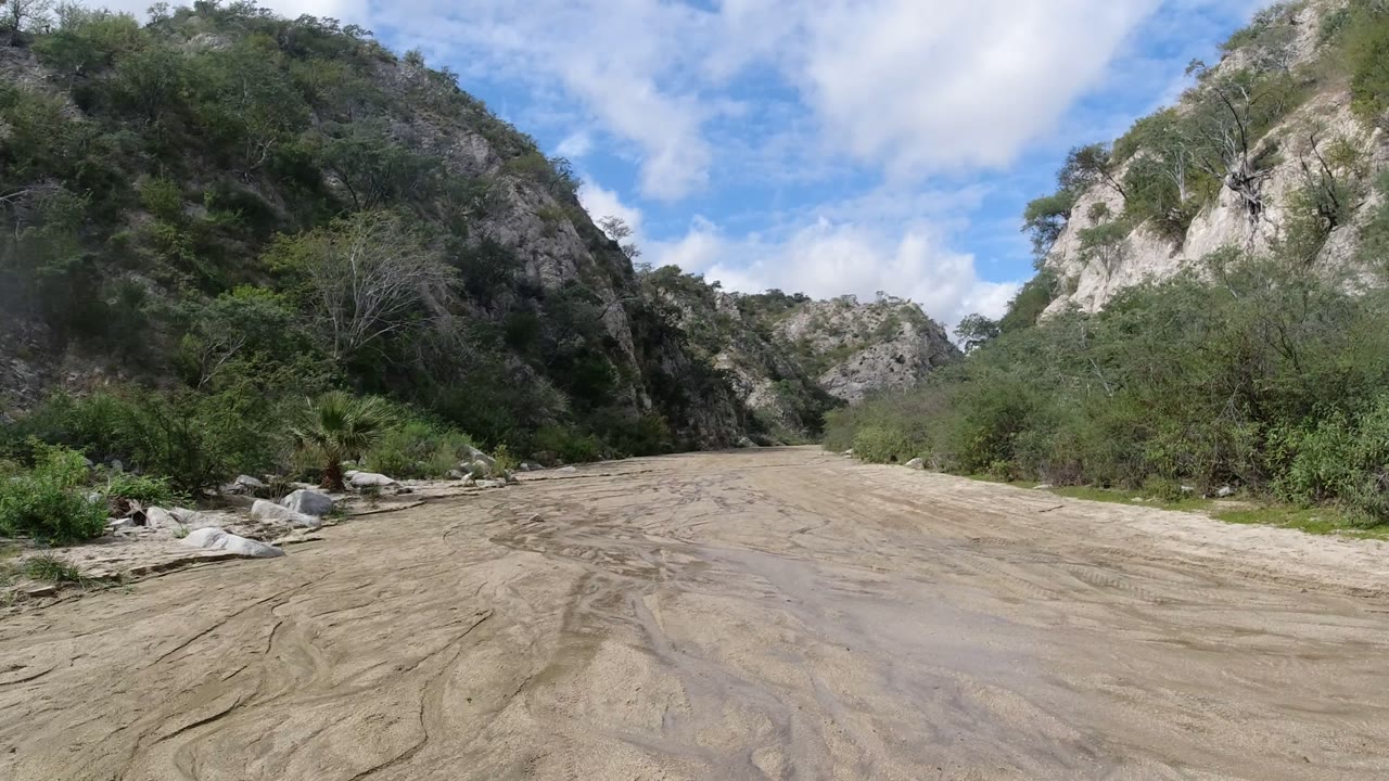 Wide ground-level view of a muddy road or riverbed between rocky green hills near San Jose del Cabo, Mexico, with shrubs at the edges and no visible people.