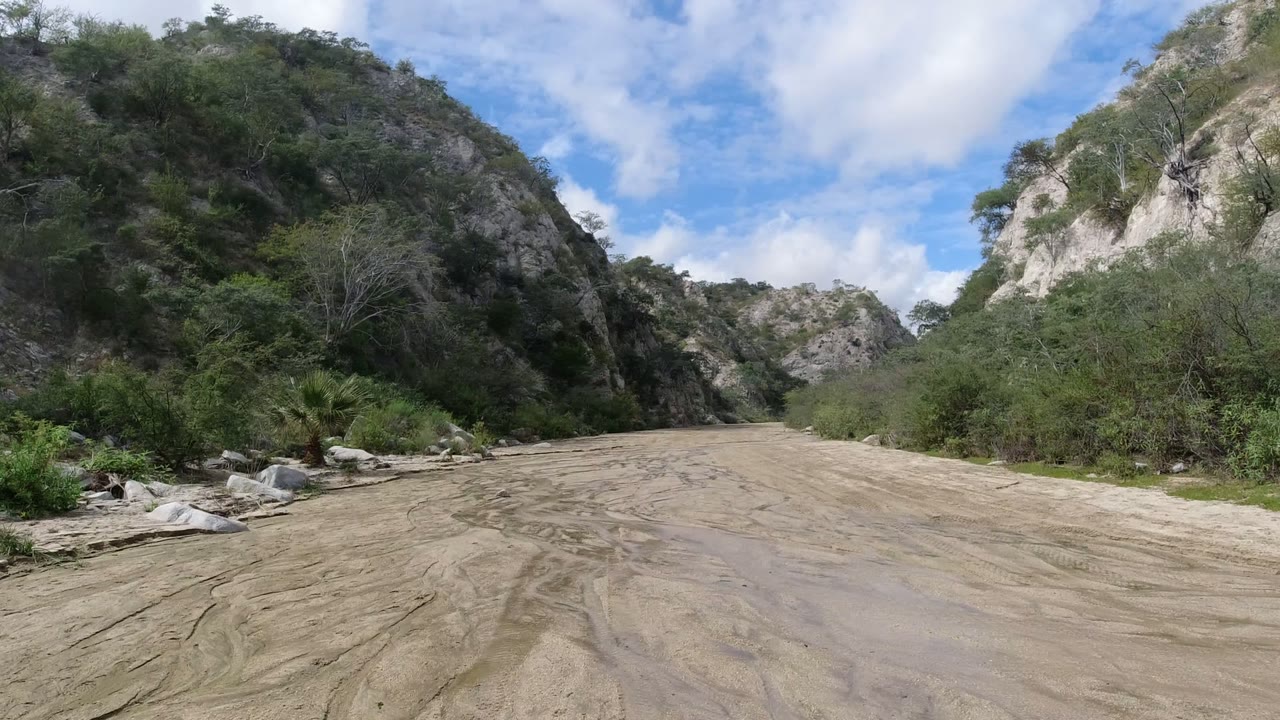 The muddy track continues through a scrub-lined valley near San Jose del Cabo, Mexico, with pale rock faces and cloud-filtered sunlight, and no visible people.