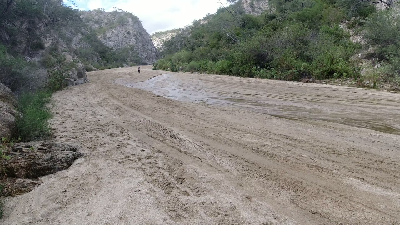 A similar wide view shows the San Jose del Cabo dirt path stretching farther into the bend, with scrub vegetation, rock faces, pale sky, and a tiny distant person near the center-left road.