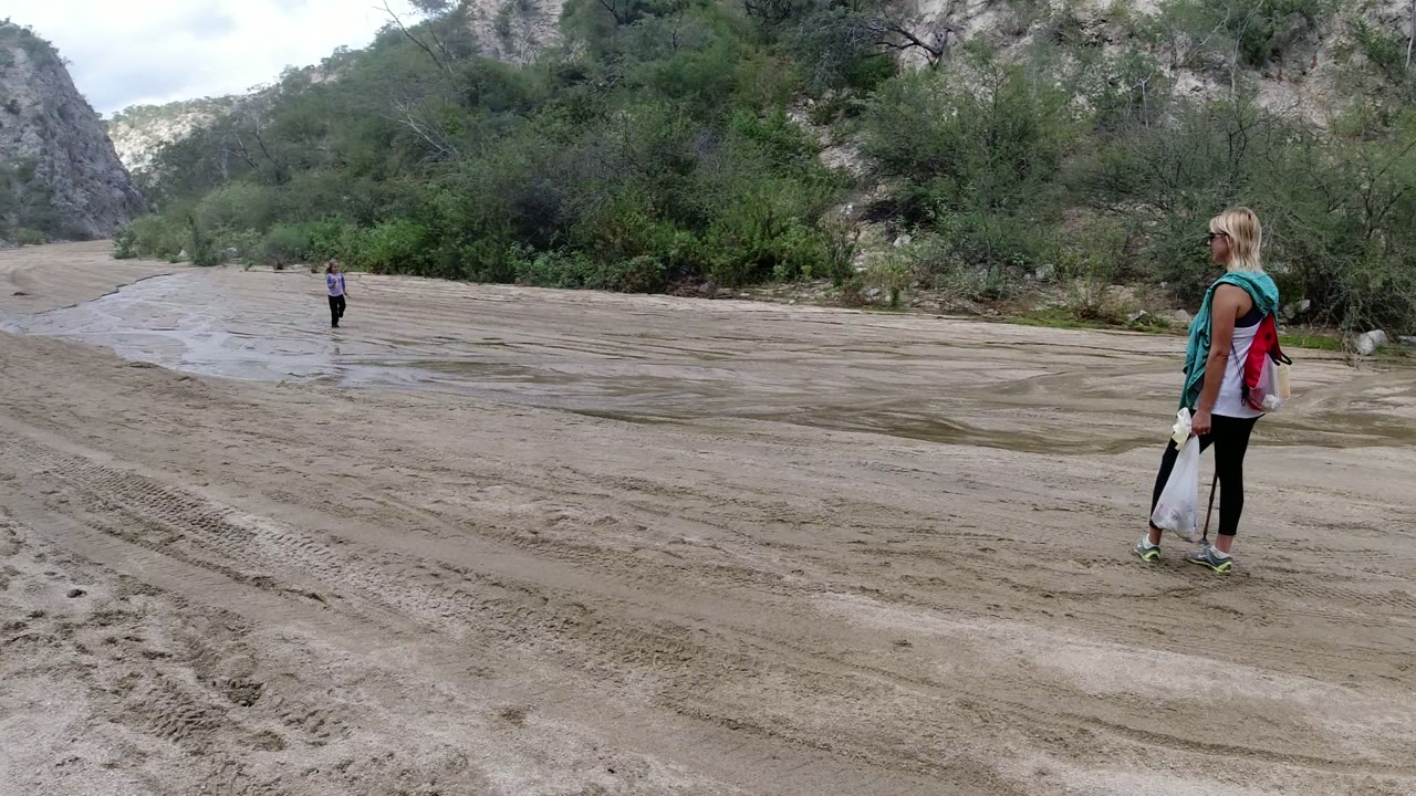 Woman watching a child on wet sand near rocky brush.