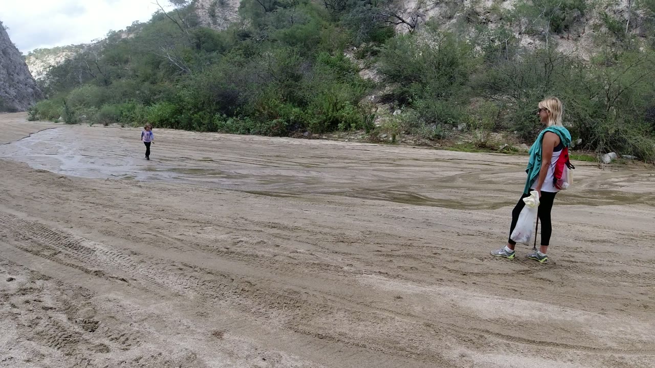 In San Jose Del Cabo, Mexico, the child remains in the open midground while the woman stands on the right side of the sandy road with vegetation and canyon rock behind them.