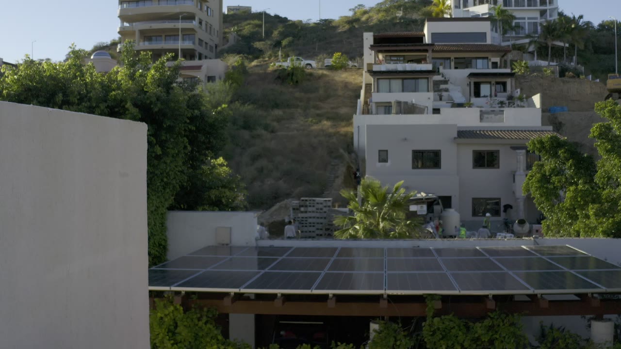 Opening frame of Los Cabos white hillside houses behind green trees, with a glass or solar-panel roof grid in the foreground and no visible people.