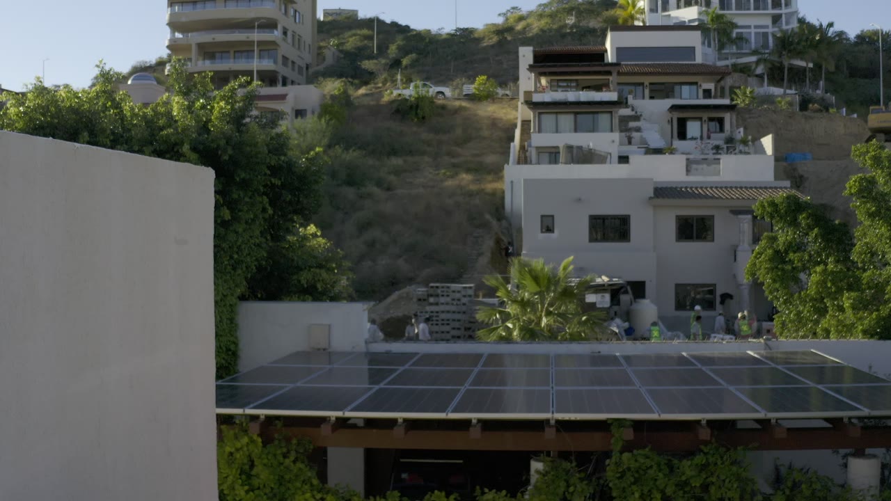 Los Cabos residential hillside frame showing a large solar-panel roof in front, light-colored duplex-style homes behind it, dry slope above, and people visible near the ground level.
