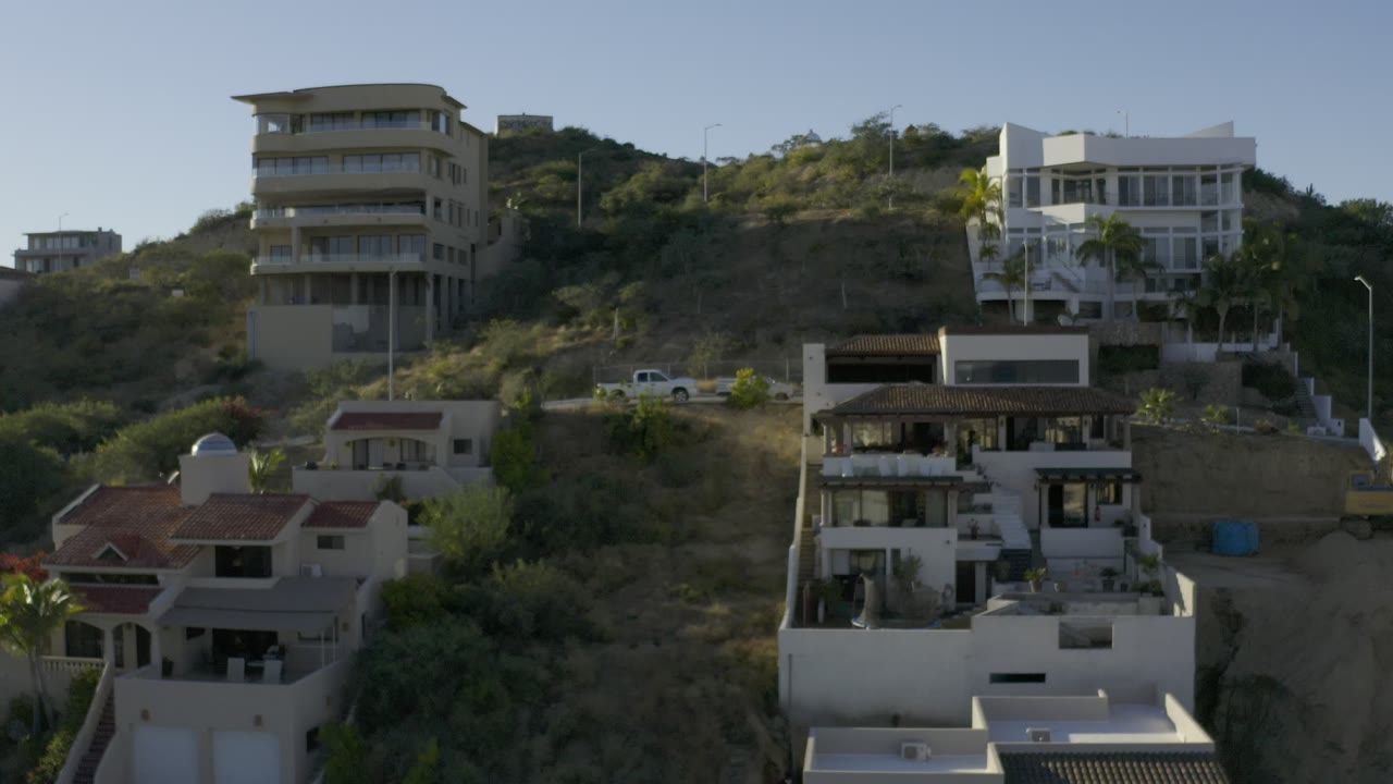 Aerial Los Cabos frame with white and tan homes stepping across a scrub-covered slope, a small vehicle on the road, and no visible people.