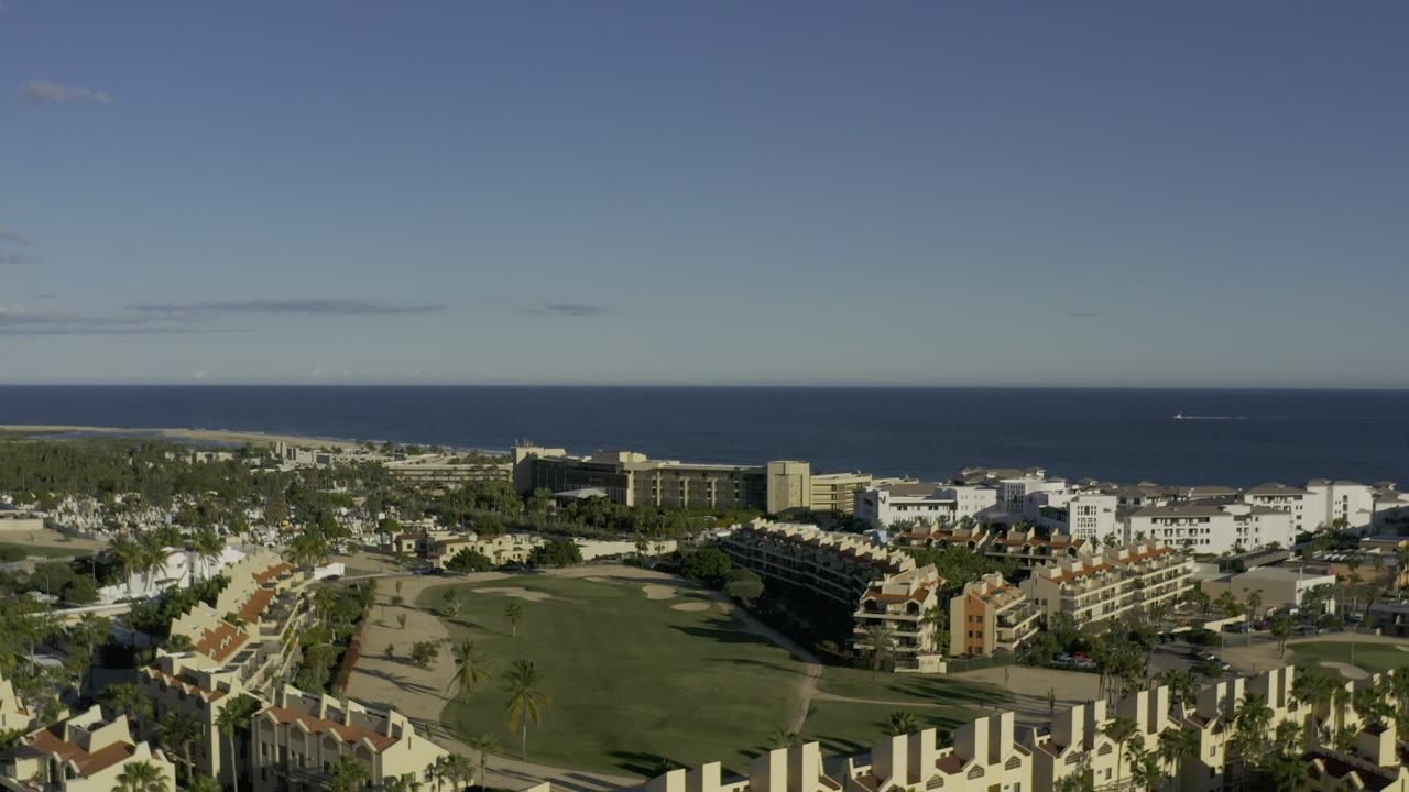 A Los Cabos, Mexico aerial frame with resort-style buildings, a green fairway-like area, palm-lined paths, and the ocean stretching across the background with no visible people.