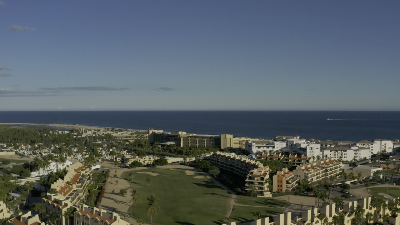 Opening aerial frame over Los Cabos, Mexico, with resort-style buildings, green grounds, palms, and a dark blue ocean horizon, no visible people.
