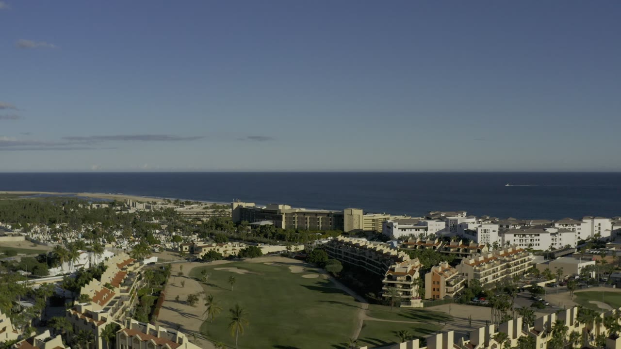 Second aerial frame in Los Cabos, Mexico, looking over landscaped lawns, clustered buildings, palms, and the ocean horizon, no visible people.