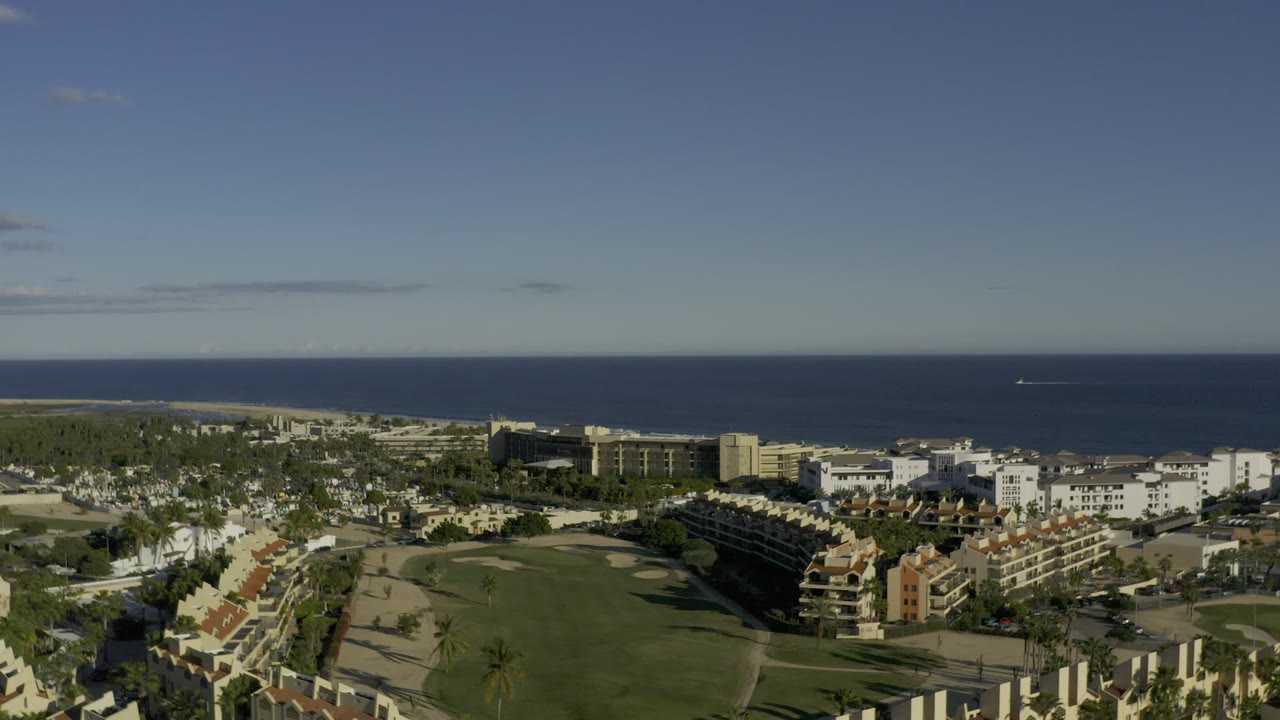 Second aerial frame in Los Cabos, Mexico, with the green central grounds and surrounding apartment-style buildings under strong daylight, with no visible people.