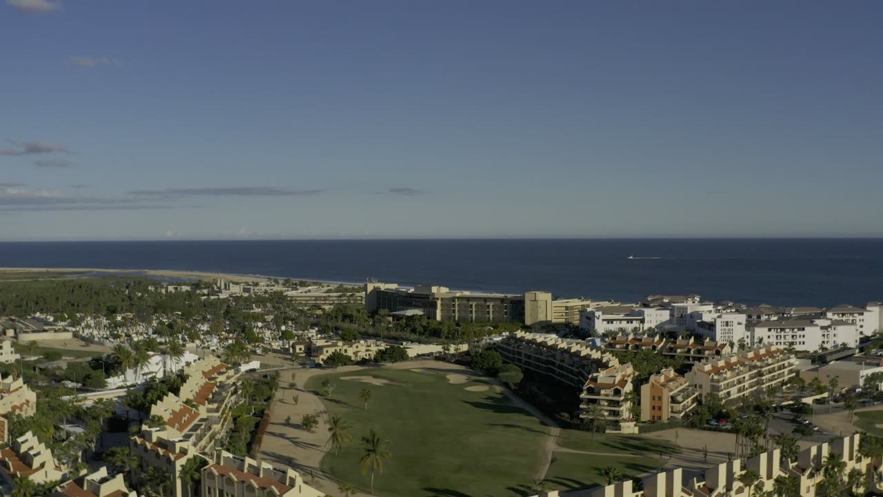 Second aerial frame in Los Cabos, Mexico, with the green resort grounds centered, rows of homes at left, hotel buildings at right, and no visible people.