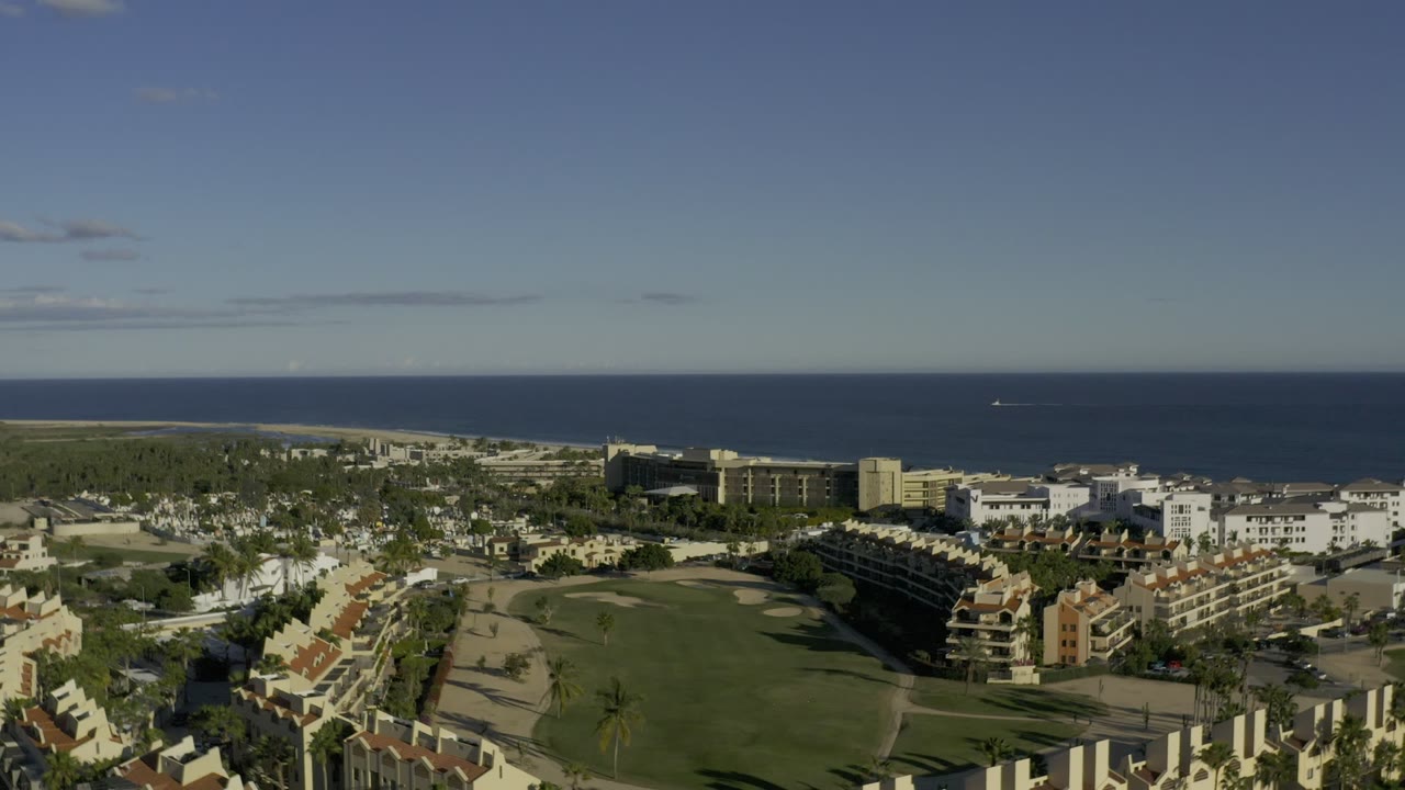 Opening frame over Los Cabos, Mexico, with low and mid-rise buildings, landscaped green areas, palms, roads, a dark blue ocean horizon, and no clearly visible people.