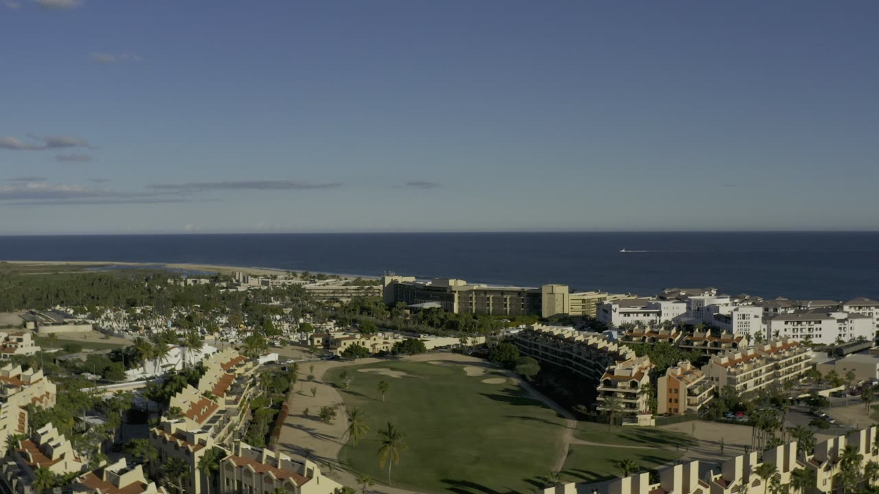 Second frame of Los Cabos, Mexico, showing spread-out buildings, a green central lawn area, palm-lined grounds, distant shoreline, and no clearly visible people.