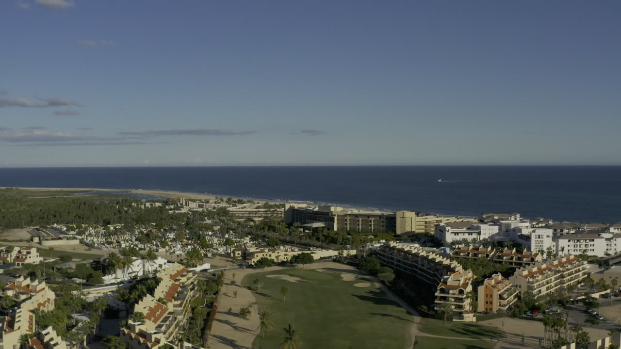 Opening aerial frame over Los Cabos, Mexico, showing low-rise residential buildings, a broad green open area, shoreline, blue ocean, and no visible people.