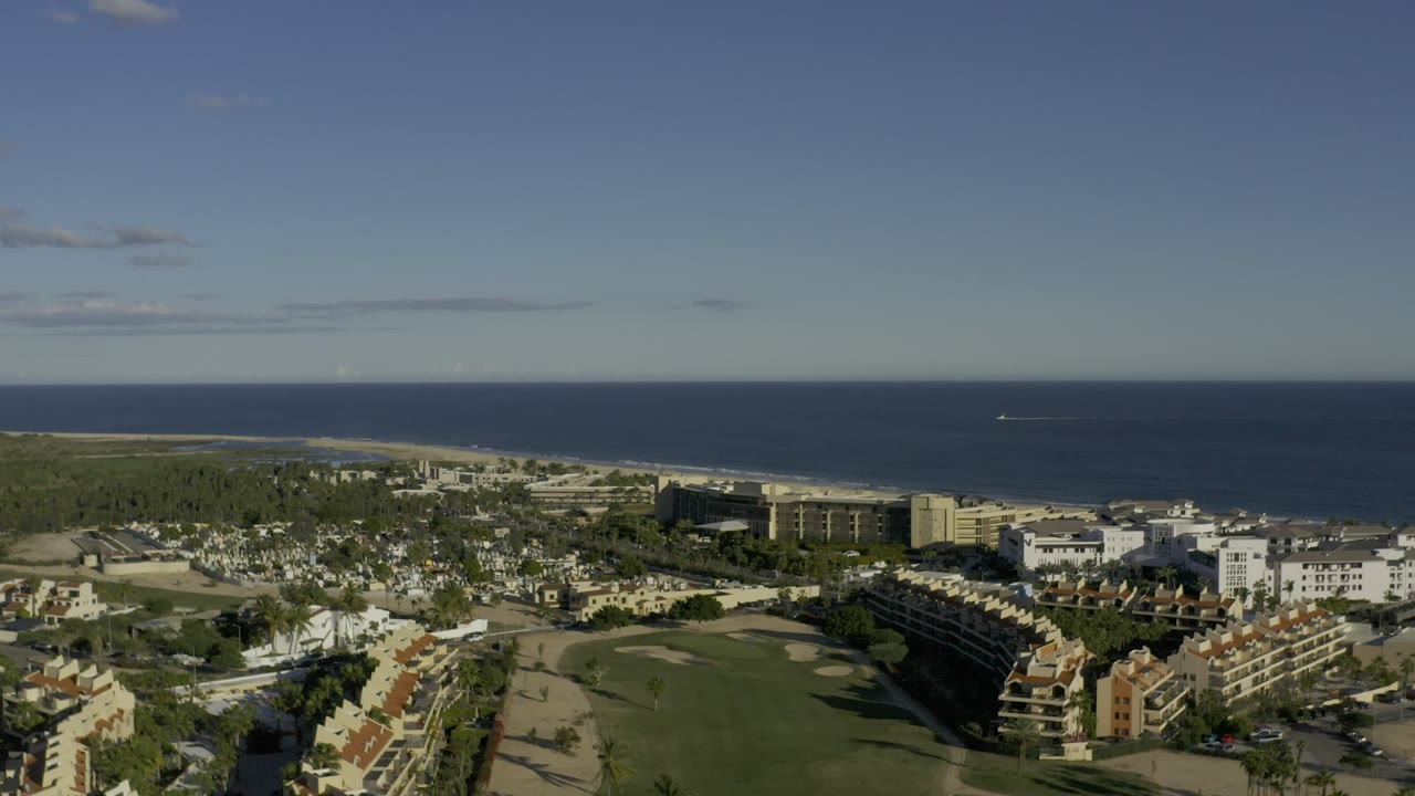 Second aerial frame in Los Cabos, Mexico, with the same coastal community slightly reframed around the green central area, distant shoreline, ocean, and no visible people.