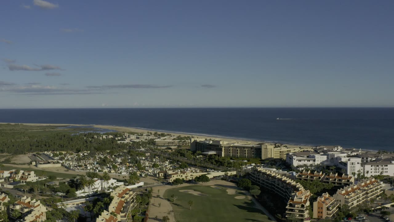 High aerial view over Los Cabos, Mexico, showing red-roofed buildings, golf course greens, sandy beach, and blue ocean horizon with no visible people.