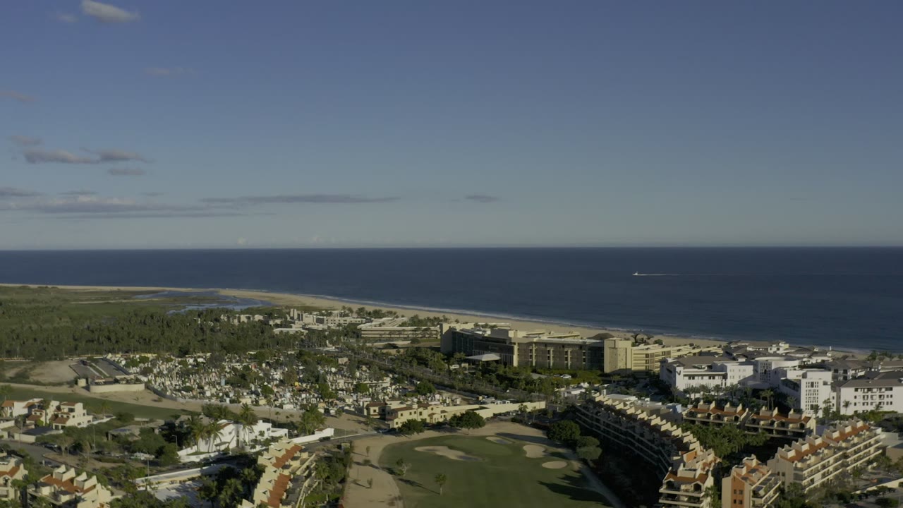 Aerial frame in Los Cabos, Mexico, with the golf course and clustered buildings below, a pale beach strip beyond, and open ocean across the horizon, with no visible people.
