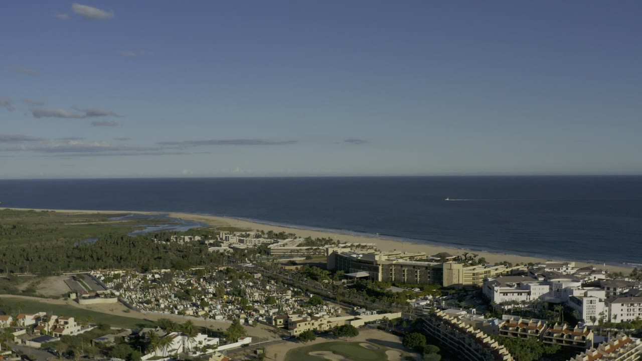 Second frame from the Los Cabos aerial view, with the shoreline running across the midground, built-up blocks below, ocean on the horizon, and no visible people.